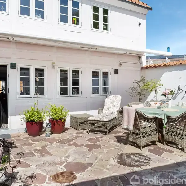 En solrig gårdhave har et spisebord med kurvestole og blomsterplanter omkring. Huset i baggrunden er hvidt med flere vinduer og en åben dør til venstre.