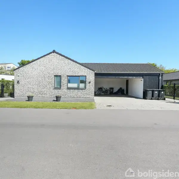 Et moderne, gråt murstenshus med fladt tag står stille på en solrig dag. På venstre side er en græsplæne, mens en carport med skraldespande er til højre.