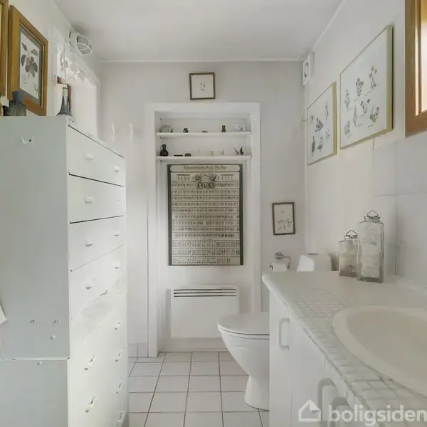 Et lyst badeværelse med hvid kommode, håndvask og toilet. Vægdekorationer består af rammer og kunst. Der hænger håndklæder, og vinduet tilføjer naturligt lys.