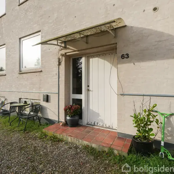 Dør markeret med tallet 63 står ved siden af et glasbord med stole. Grusbelagt indkørsel og planter i krukker supplerer ydersiden af et hvidt murstenshus.