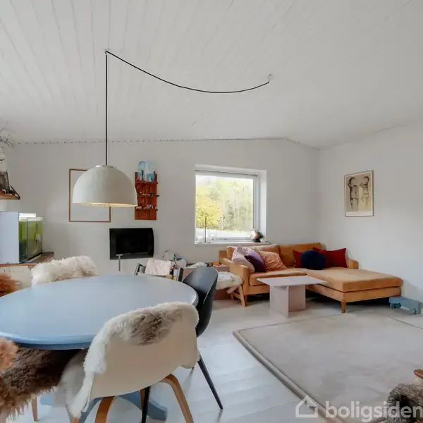 Rundt spisebord med pelsklædte stole står i stue med lyse vægge. Sofaområde med farverige puder ses i hjørnet. Vinduer giver naturligt lys. Møbler og dekoration skaber en hyggelig atmosfære.