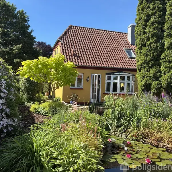 Gult hus med rødt tegltag omgivet af frodig have med mange grønne planter og trær. En lille dam i forgrunden med nærliggende blomster skaber en idyllisk atmosfære.