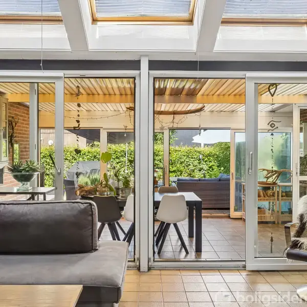 Stue med grå sofa, hvor sollys strømmer ind gennem ovenlysvinduer. Skydedøre åbner ud til en terrasse med spisebord og stole, omgivet af grønne planter og plankeværk.