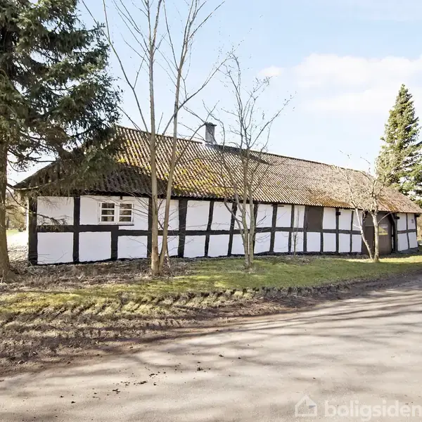 Bindingsværkshus står stille omgivet af grusvej, bartræer og græsareal. Husets hvide vægge med sorte bjælker kontrasterer med den brunlige tagflade under en skyfri himmel.