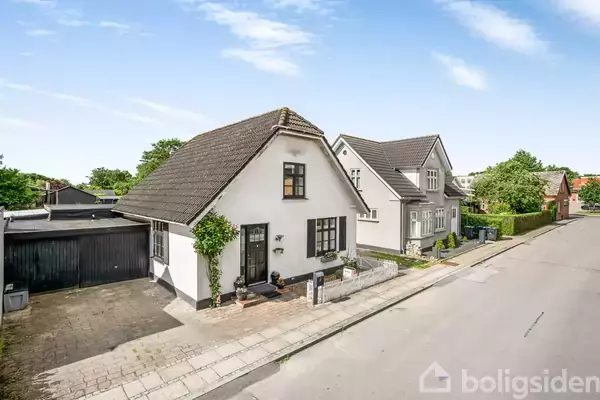 Et hvidt hus med skråt sort tag står langs en stille gade. Ved siden af huset er en carport, og der er grønt løv omkring. Naboen har et lignende, men større hus.