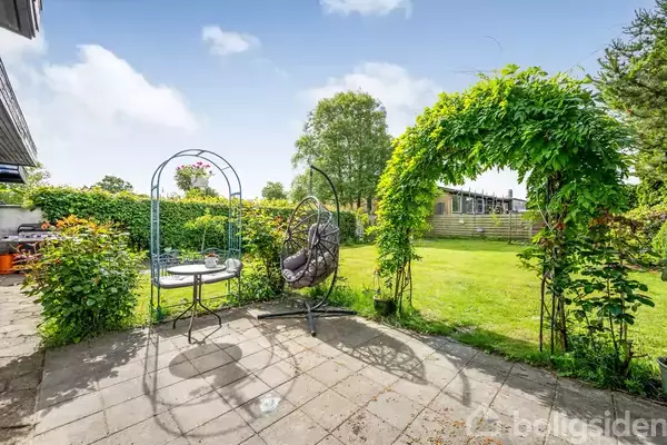 En hængestol og et cafebord med to stole står på en flisebelagt terrasse. En bue med grønne klatreplanter fører til en velplejet græsplæne i baghaven.