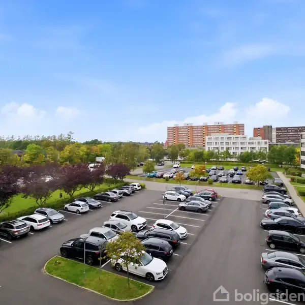 Flere biler holder parkeret på en stor asfalteret parkeringsplads. I baggrunden ses grønne træer og flere boligbygninger under en blå himmel.