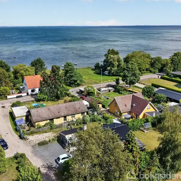 Flere huse ligger nær kysten ved en rolig, skovrig baggrund. De er omgivet af grønne haver og træer. Kystlinjen strækker sig langs en stor vandflade i baggrunden.