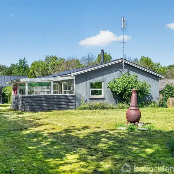 Et gråt hus med store vinduer står i en frodig have. Huset har en antenne på taget. Plænen er velplejet, omgivet af træer, et drivhus og en rød udendørs pejs.