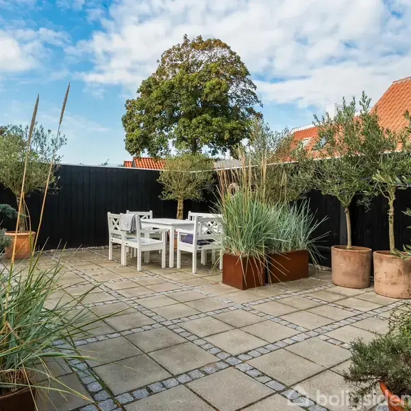 En terrasse med hvide udemøbler placeret på et flisebelagt område. Omgivet af planter i krukker med træer og buske. Solrøde tagtage og blå himmel i baggrunden.