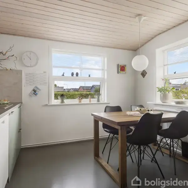 Et lyst køkken med hvidt loft og vægge, grå gulv, har et træbord med sorte stole. Planter og dekorationer står i vinduet. Et ur og køkkenredskaber hænger på væggen.