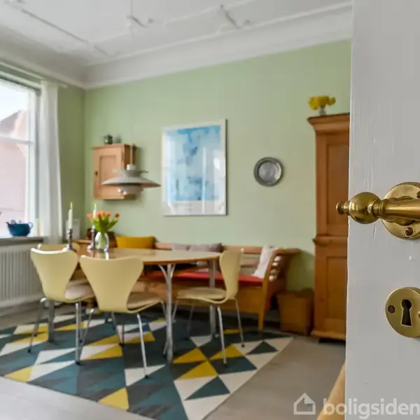 Dørhåndtag i messing åbner ind til spisestue. Fire stole og bord står på geometrisk gulvtæppe, omgivet af grøn væg, stor vindueskarm med planter, skab og vægdekorationer.