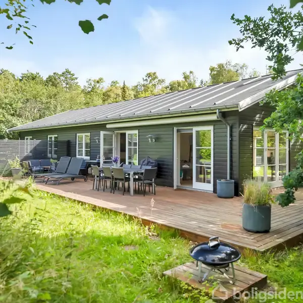 Træhus med stor træterrasse udstyret med havemøbler og grill placeret i en frodig have omgivet af skov og buske. Solskinsvejr giver en lys og indbydende atmosfære.