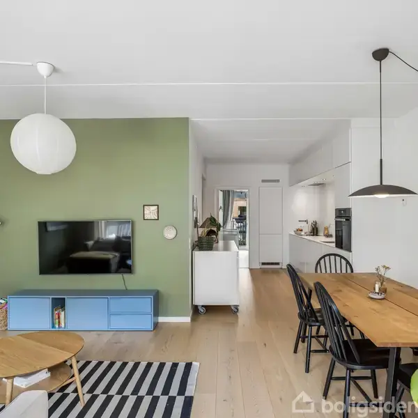 En stue med TV på grøn væg og blå skænk. Træbord og stole til højre, stribet tæppe på gulvet. Moderne køkken i baggrunden. Vægdekorationer og planter.