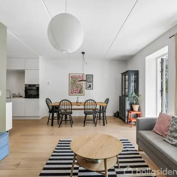 Stue med moderne møbler; en sofa, sofabord og TV i forgrunden. Et spisebord med stole står i baggrunden. Rummet er lyst med store vinduer og en grøn plante.