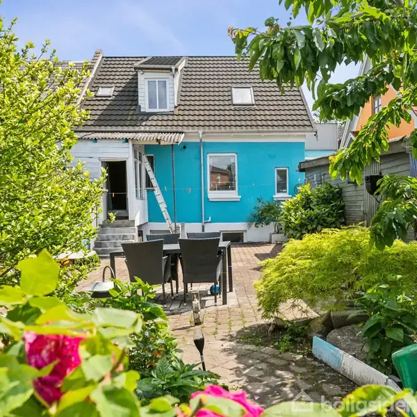 Huset er malet i en lyseblå farve og har en stige op ad siden. I forgrunden står et sort havebord med stole på en brolagt terrasse, omgivet af frodig have.