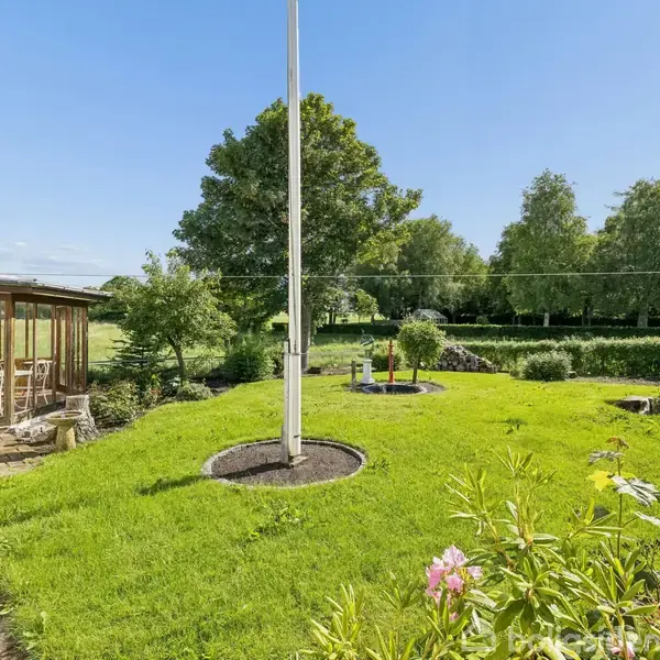 Flagstang står i en velholdt have med grøn græsplæne. Ved siden af er en terrasse med glasinddækning, indeholdende hvide møbler. Baggrunden består af træer under en klar blå himmel.