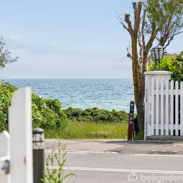 Hvid havelåge står åben og peger mod havet. Haven er omgivet af grønne buske og træer, og huset ligger ved siden af indgangen.