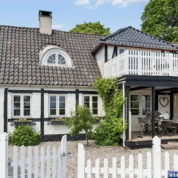 Hus med sort tegltag og hvid facade har terrasse med møbler. Omgivet af hvidt stakit og grønne planter. Skilt viser "234 B".