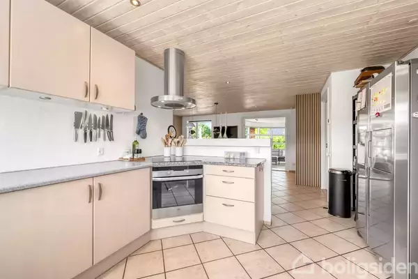 Et moderne køkken med en indbygget ovn og kogeplade, omgivet af lyse skabe. Knive hænger på væggen. I baggrunden ses en spiseplads med grønt udenfor vinduerne.