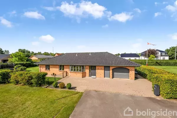 En rødstensvilla med sort tegltag står på en velplejet græsplæne med indkørsel, omgivet af buske og hække, under en klar blå himmel. En flagstang ses til højre.