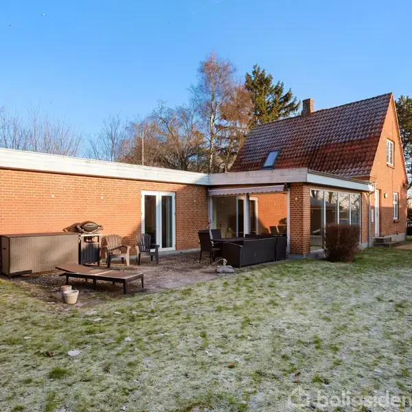 En rød murstensvilla står i en have med terrasse og havemøbler. Græsset er let frostdækket, og der er træer i baggrunden.