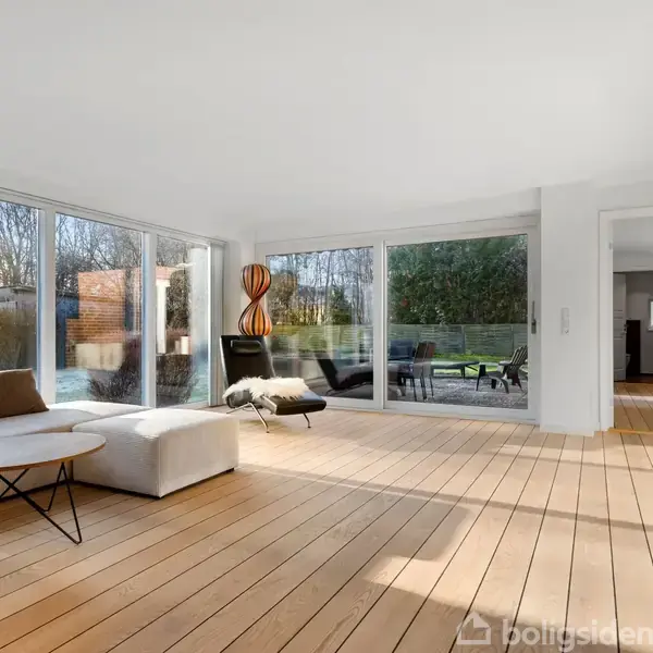 Lys stue med en hvid sofa og rundt bord, badet i sollys fra store glasvinduer, åbner ud til en terrasse og have.