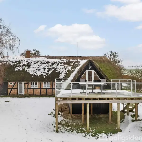 Et bindingsværkshus med stråtag er dækket af let sne. Huset har en stor træterrasse med glasrækværk. Det omkringliggende landskab er også sneklædt, og himlen er let overskyet.