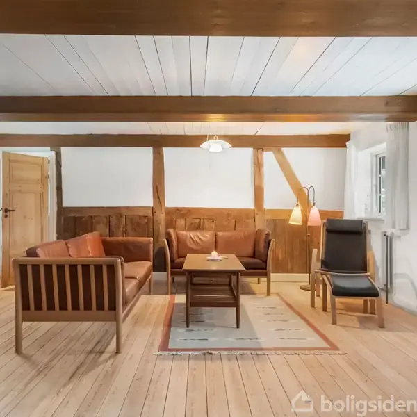 En stue med træmøbler står stille i et hyggeligt rum med synlige loftsbjælker. To sofaer og en lænestol omkranser et sofabord på et tæppe. Væggene har træpaneler.