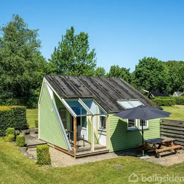 Et lille, lysegrønt sommerhus med skråt tag står på en velplejet græsplæne. En parasol dækker et picnicbord i solrig have omgivet af buske og træer.