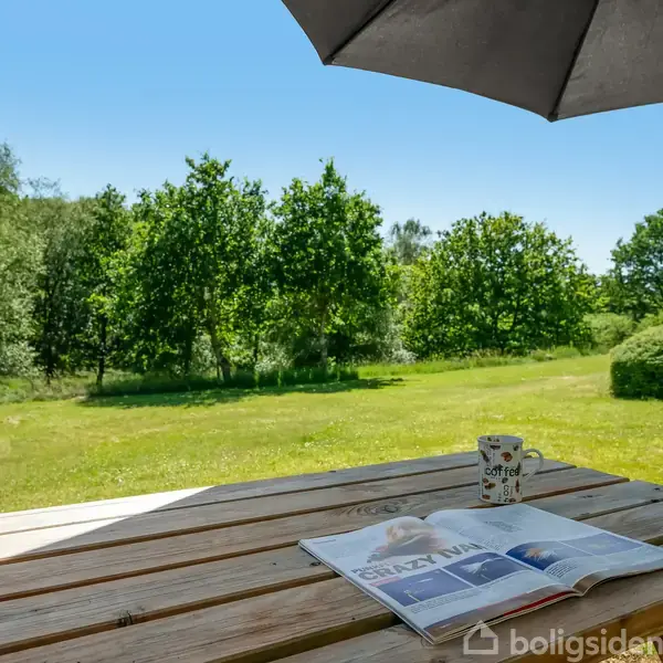 Et træbord står under en parasol. En kop og en åben avis ligger på bordet. Omgivet af en grøn græsplæne og træer på en solrig dag.