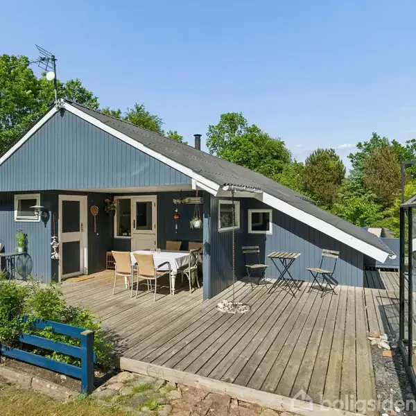 Et blåt sommerhus med asymmetrisk tag står på en træterrasse. Et bord med stole er placeret ved huset, omgivet af grønne træer og buske. Ingen tekst til stede.