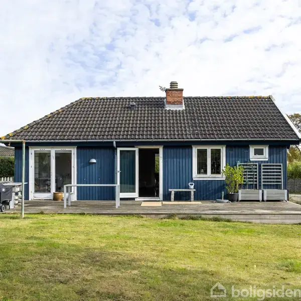 Et blåt træhus med sorte tegltag står på en plæne. Huset har vinduer og en dør, der fører ud til en terrasse. Græsplænen omgiver bygningen.