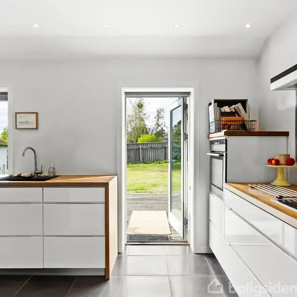 Et moderne køkken med hvide skabe og træborde. En dør åbner ud til en græsplæne og hegn. En kogeplade, håndvask, og ovn er synlige.
