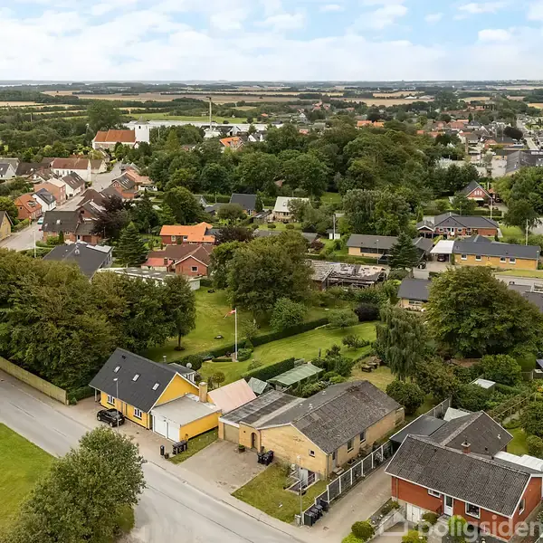 Bebyggelsens huse står stille i en grøn forstadsomgivelser med træer og marker i horisonten under en delvist skyet himmel.