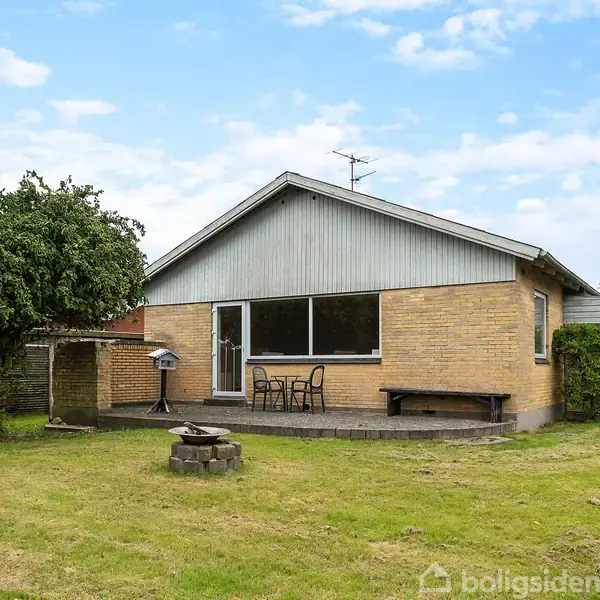Et enkelt etplanshus med murstensvægge og et græsareal foran. Der er en terrasse med borde og stole, omgivet af træer og buske under en blå himmel.