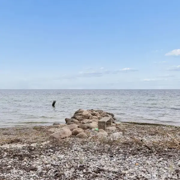 Strand med sten og tangstriber, midtpunktet er en stenhøj. Havet breder sig i baggrunden under en klar blå himmel, en person ses i vandkanten. Ingen tekst til stede.