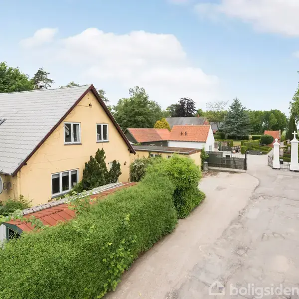 Et gult hus med rødt tag står stille omgivet af grønne buske og træer, ved siden af en bred indkørsel og en hvid port i et roligt, grønt boligkvarter.