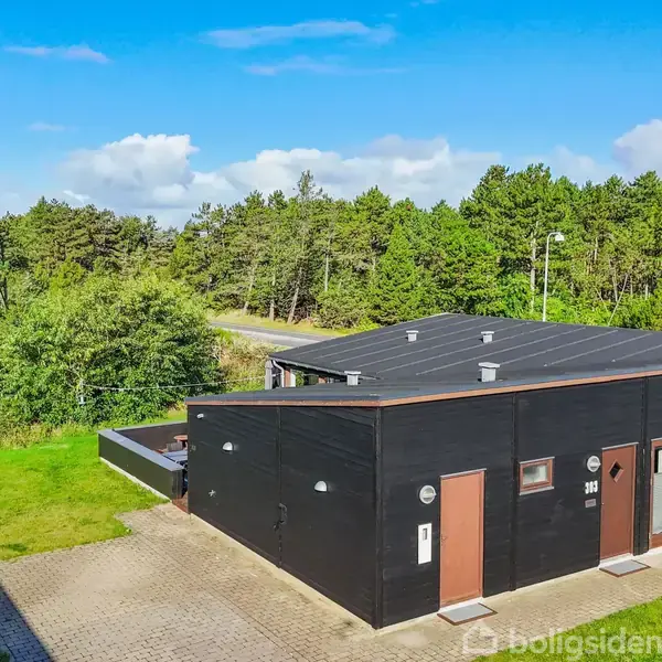 En sort, firkantet bygning med fladt tag står på en brolagt terrasse. Omkringliggende træer og græsplæne skaber en naturskøn baggrund under en klar blå himmel.