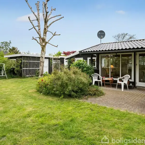 Huset har en udendørs terrasse med hvide stole og et bord. I haven er der et græsområde, en lille busk og et træ; haven er omgivet af buske.