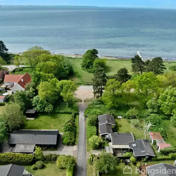 Kystnære sommerhuse ligger blandt træer, med en sti der fører til en strand ved havet. Grønt landskab omgiver området med enkelte huse, tæt på vandkanten.