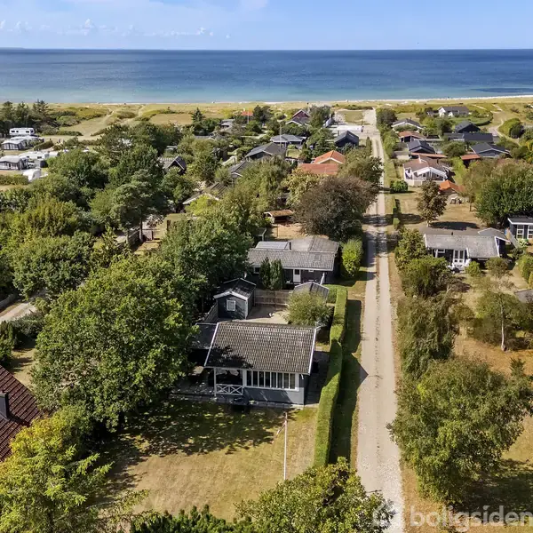 Et sommerhusområde ligger tæt ved kysten. Huse omgivet af træer strækker sig langs en vej med udsigt til havet. Solrig dag med få skyer og klart blåt hav i baggrunden.