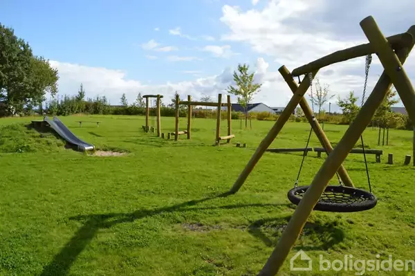 Legeplads med gynge og rutsjebane står tom i en grøn park omgivet af træer under en blå himmel med skyer. Text: "boligsiden".