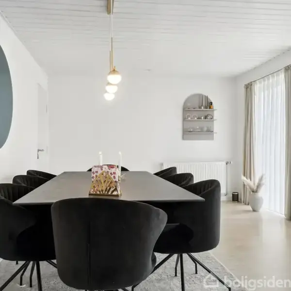 Et elegant spisebord omgivet af sorte stole står i et lyst rum. To stearinlys og en servietstander er på bordet. En rund spejl hænger på væggen; store vinduer lader lys ind.