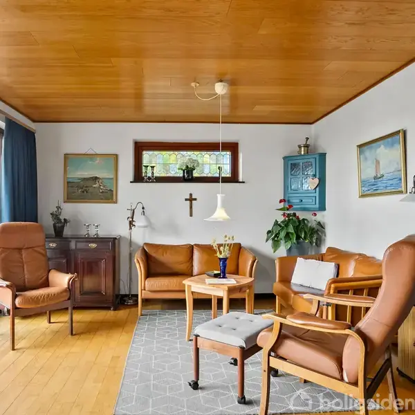Brunt møblement står i en stue med trægulve og træloft. Sofa og stole omkranser et lille bord. Væggene har malerier, et kors, og en indbygget reol. Dobbeltdøre fører udenfor.