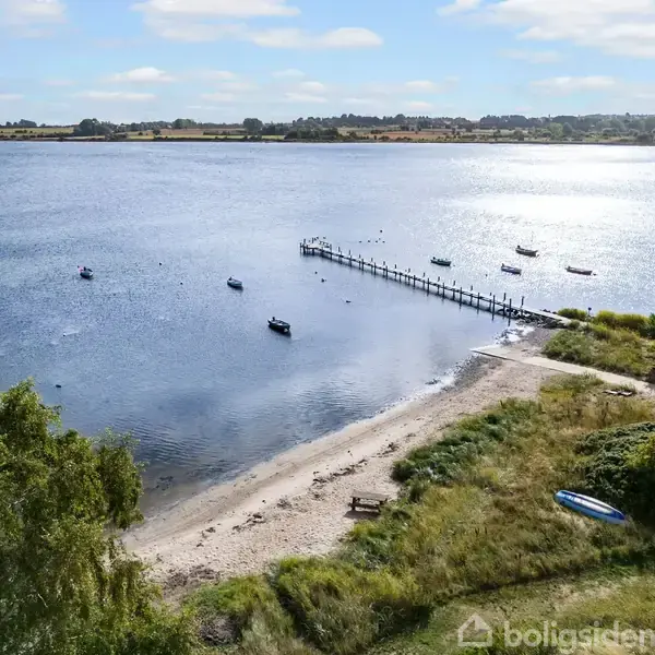 En strand med en træbro strækker sig ud i en rolig sø med flere små både. Omkring er der græsarealer og buske under en klar blå himmel.