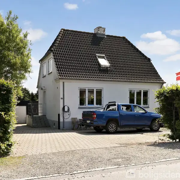 Et hvidt to-etagers hus har en blå pickup parkeret foran. I baggrunden er en grøn hæk og et træ. Det danske flag ses ved siden af huset.