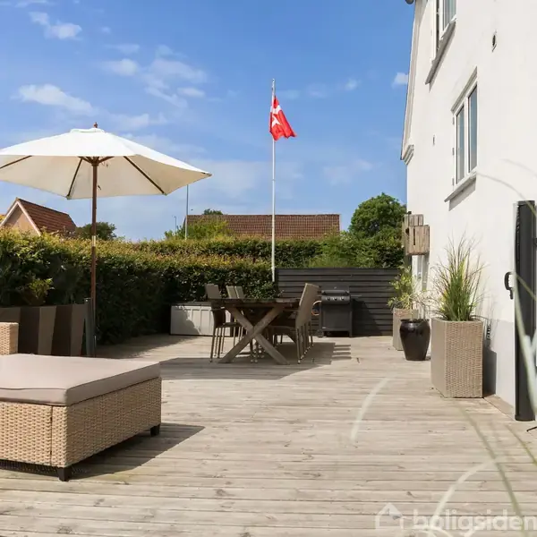 Terrasse med siddemøbler og parasol i sollys, omgivet af hække og blomsterkrukker foran et hus. Et bord er opstillet nær et dansk flag.