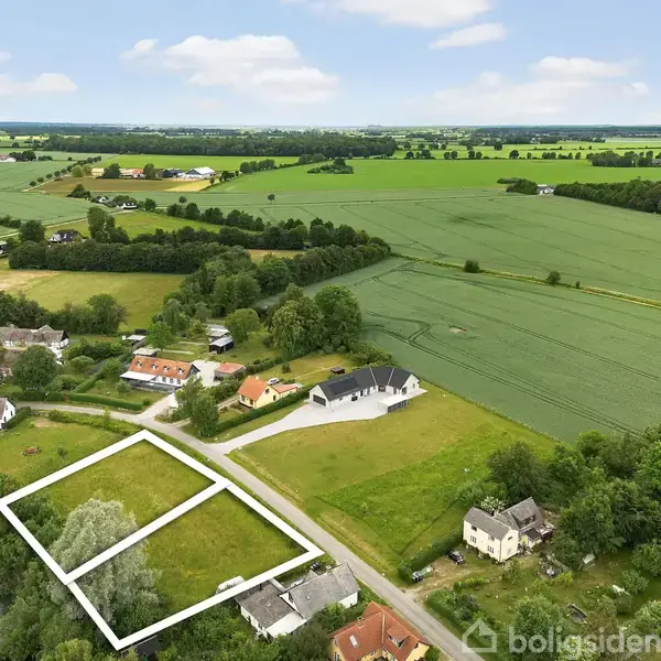 En grunde markeret med hvide linjer ligger ved en landevej omgivet af marker og huse tæt på en sø. Landskabet er frodigt og landligt med marker i baggrunden.