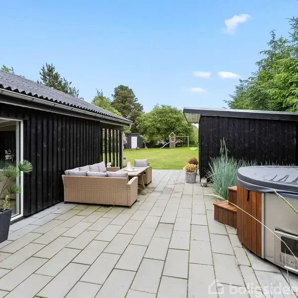 Udendørs terrasseområde med brune loungemøbler og en rund spa-bad på flisebelagt terrasse. Omgivet af træfacader, grønne planter og en græsplæne i baggrunden under en blå himmel.